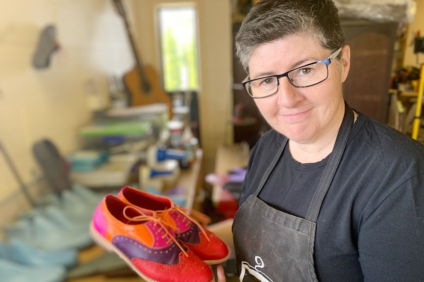 A woman with glasses holds a par of orange and pink handmade shoes