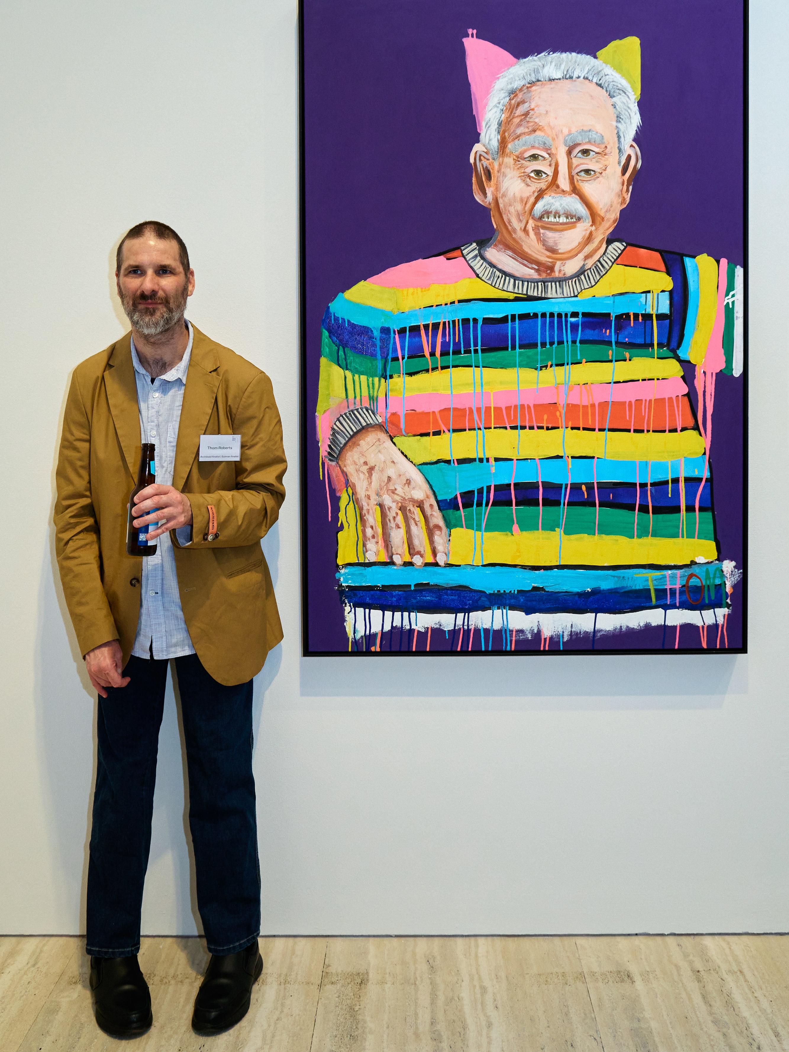 Artist wears a brown suit jacket, standing next to his brightly coloured painting of Ken Done, in Art Gallery NSW