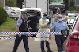 Police and health workers wear PPE as they walk behind police tape in a street.