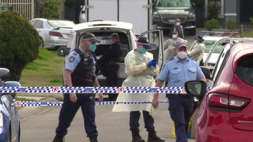 Police and health workers wear PPE as they walk behind police tape in a street.