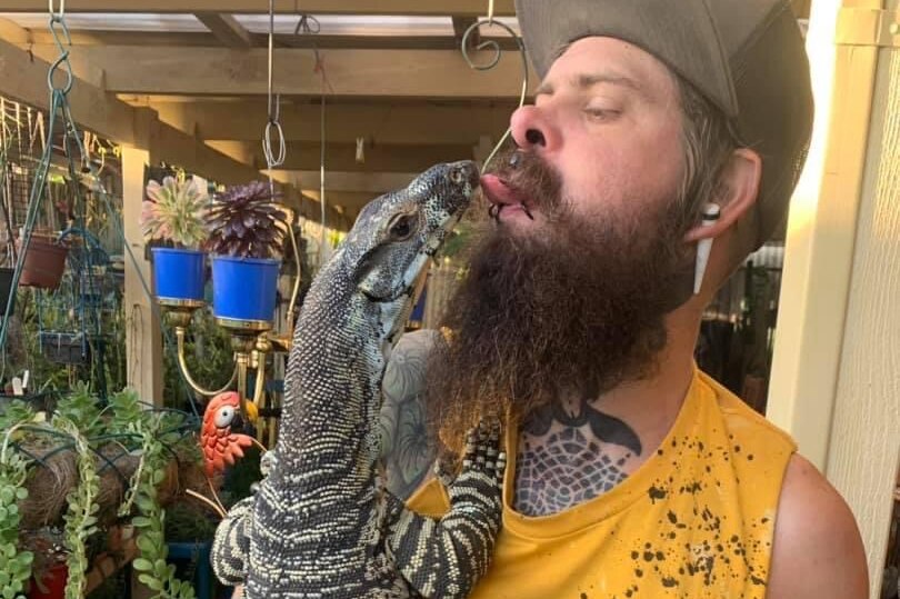 A tattooed man cuddling a large lizard gives it a lick on the snout.