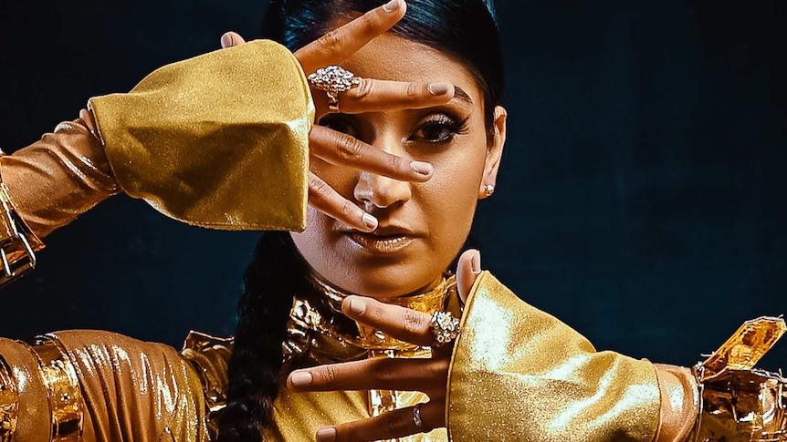 Parvyn wears a gold outfit with gold jewellery. She is looking directly at the camera with her hands in front of her face.