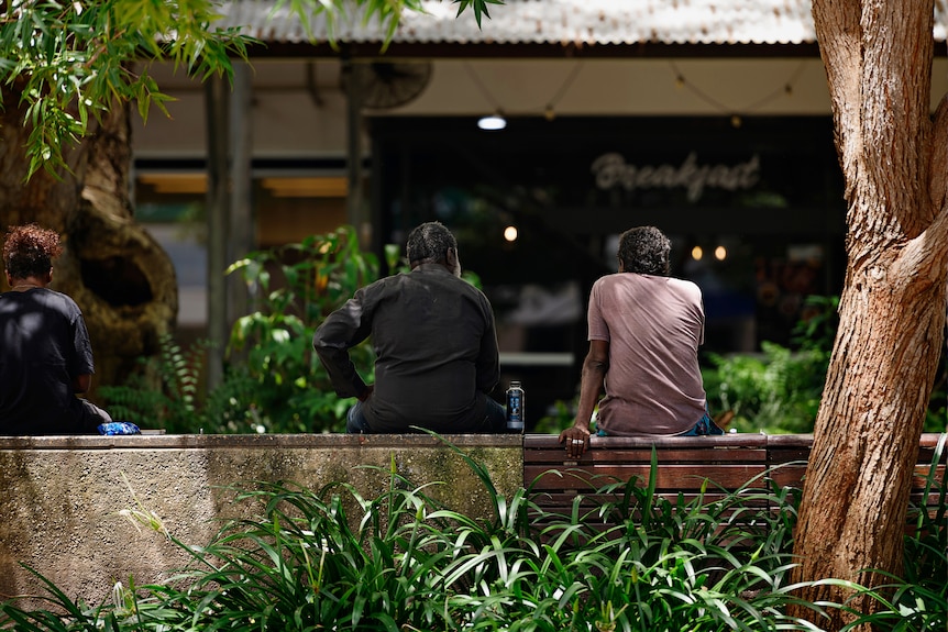 Two Indigenous people sit on a bench outside in Darwin, they face away from  the camera.