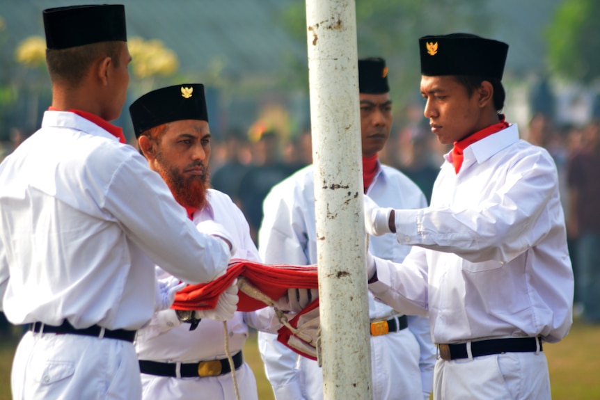 Umar Patek dressed in a white formal-looking uniform raising a flag with three other men. 