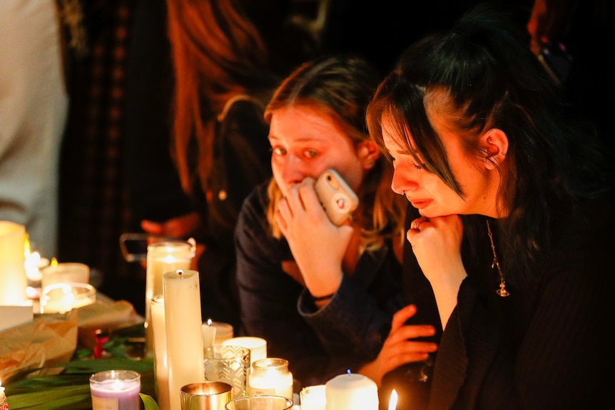 Two women lean over a table lit with candles as they shed tears