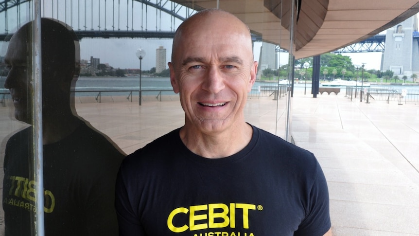 Stephen Scheeler, wearing a black t-shirt in front of a glass wall, reflecting the Sydney Harbour Bridge.