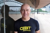 Stephen Scheeler, wearing a black t-shirt in front of a glass wall, reflecting the Sydney Harbour Bridge.