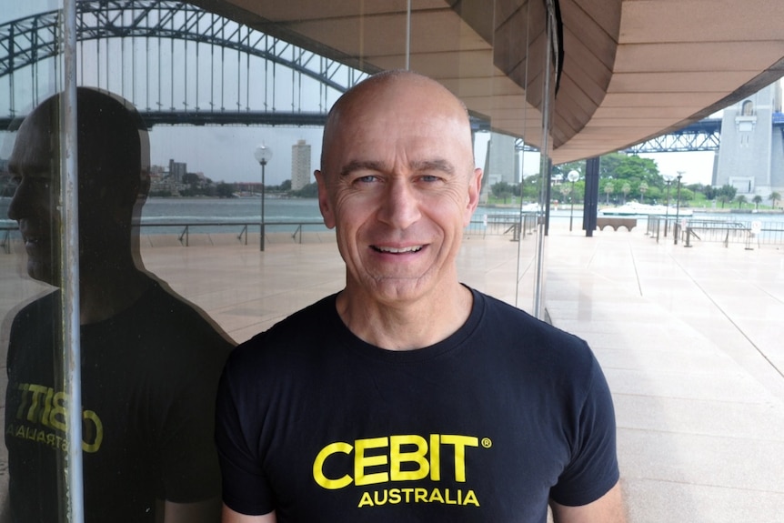 Stephen Scheeler, wearing a black t-shirt in front of a glass wall, reflecting the Sydney Harbour Bridge.