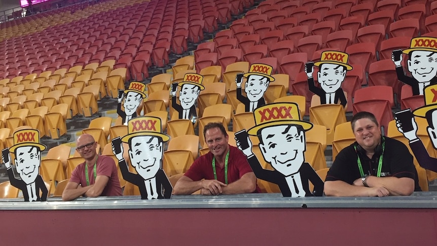 Three men sit in an empty stand surrounded by cut outs of a black and white picture of a winking man with a yellow hat on