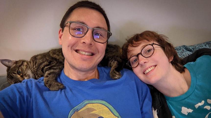 Two people sitting with their heads close and a tabby cat drapped across their necks