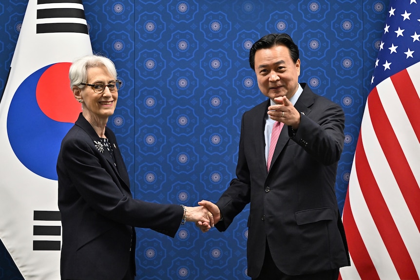 Două persoane își strâng mâna în fața drapelului Coreei de Sud și Statelor Unite.