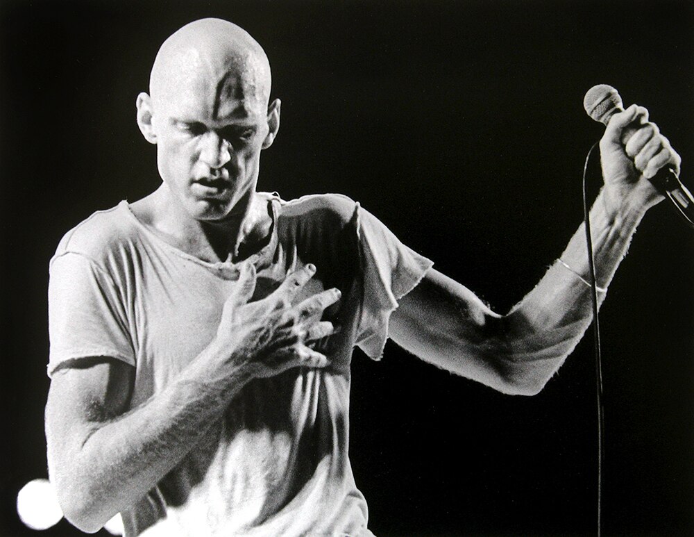 A bald man holds a microphone in one hand and holds his other hand over his heart