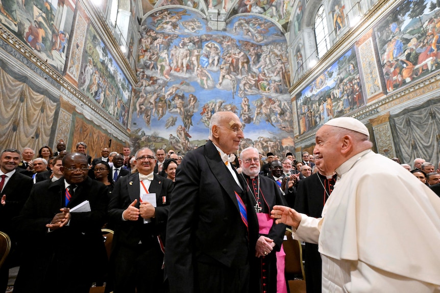 An elderly man in a white cap and gown speaks to another elderly man in a black suit in a Vatican hall covered in murals.
