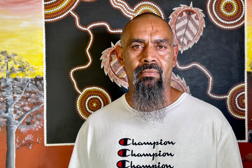 Indigenous man with a beard wearing a white Champion t-shirt standing in front of traditional art.