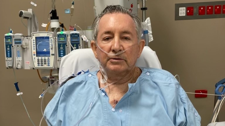 70-year-old man in blue gown sitting up in a hospital bed. 