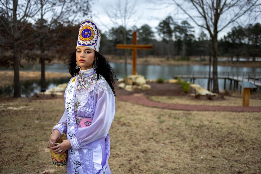 Shemah Crosby in a purple and white dress and headdress looking toward the camera with a cross and lake in the background