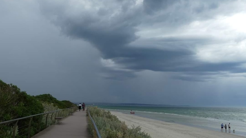 Dark storm clouds gather over a beach in Victoria as people walk along the sand.