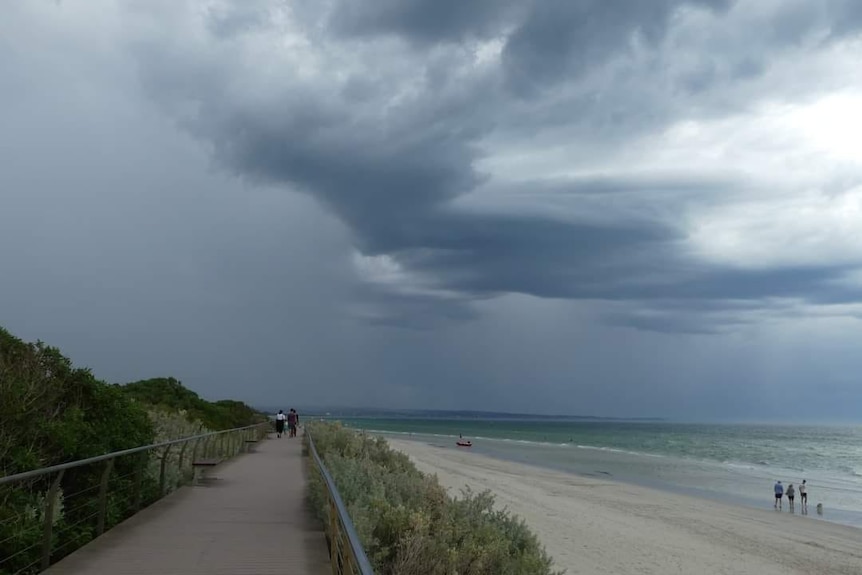 Dark storm clouds gather over a beach in Victoria as people walk along the sand.