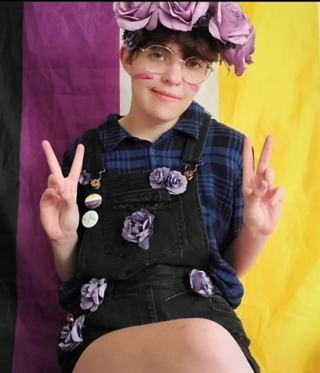 A person wearing a crown of roses sitting in front of a purple, white and yellow flag doing peace signs