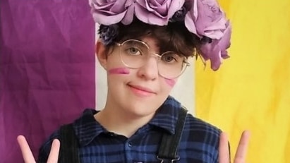 A person wearing a crown of roses sitting in front of a purple, white and yellow flag doing peace signs