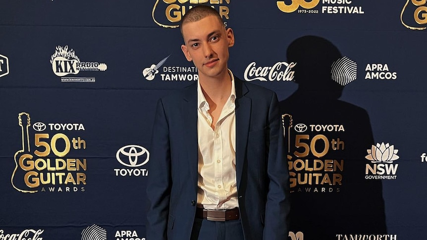 Young man in a suit smiles and stands in front of Golden Guitar media wall