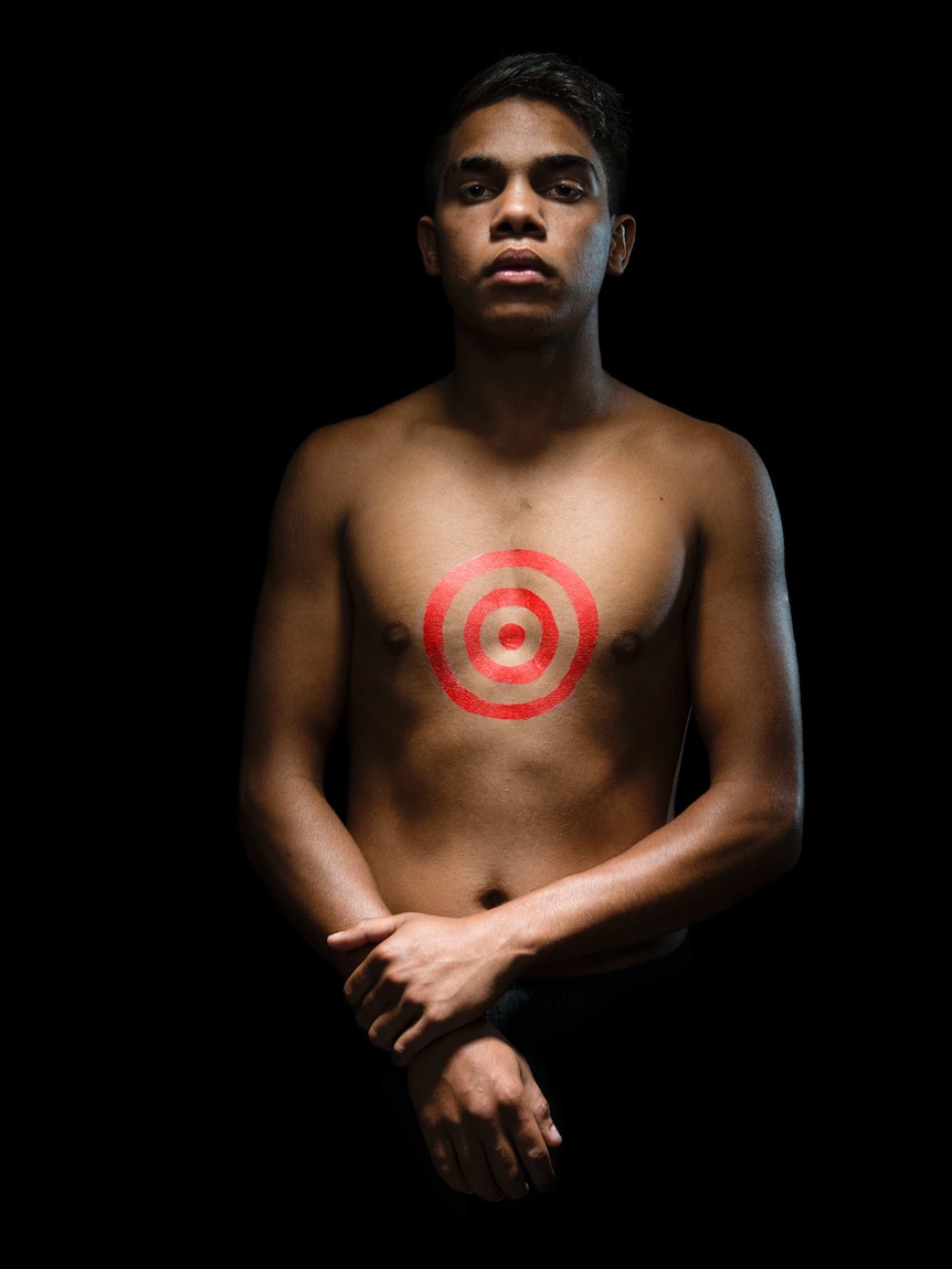 A young man stands topless against a black background, lit by warm light, with a red target symbol painted on his chest.