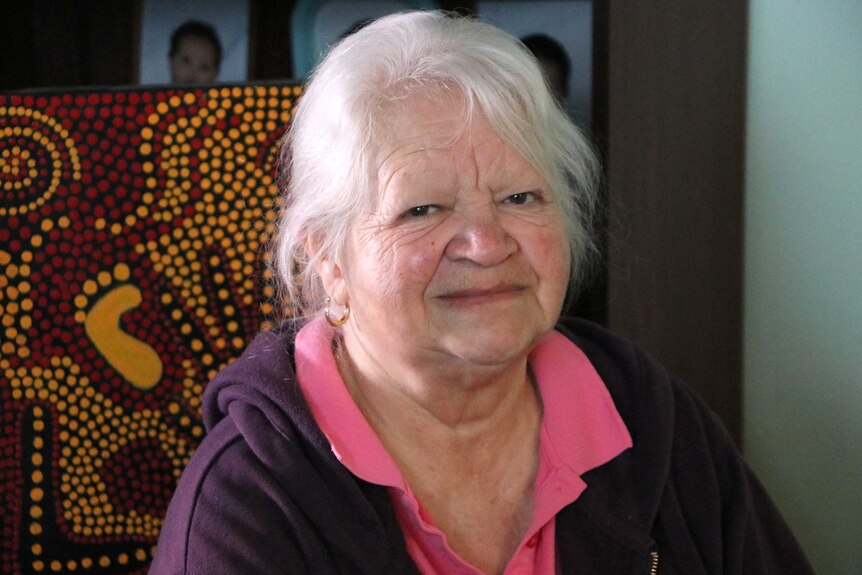 Louise sits in front of an Aboriginal dot painting with pictures of babies in the background.