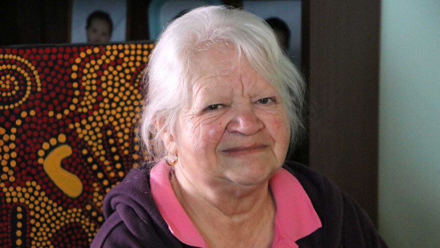 Louise sits in front of an Aboriginal dot painting with pictures of babies in the background.