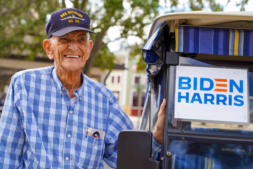 An older man in a USS Missouri navy cap next to his golf cart with a Biden-Harris sign in the window