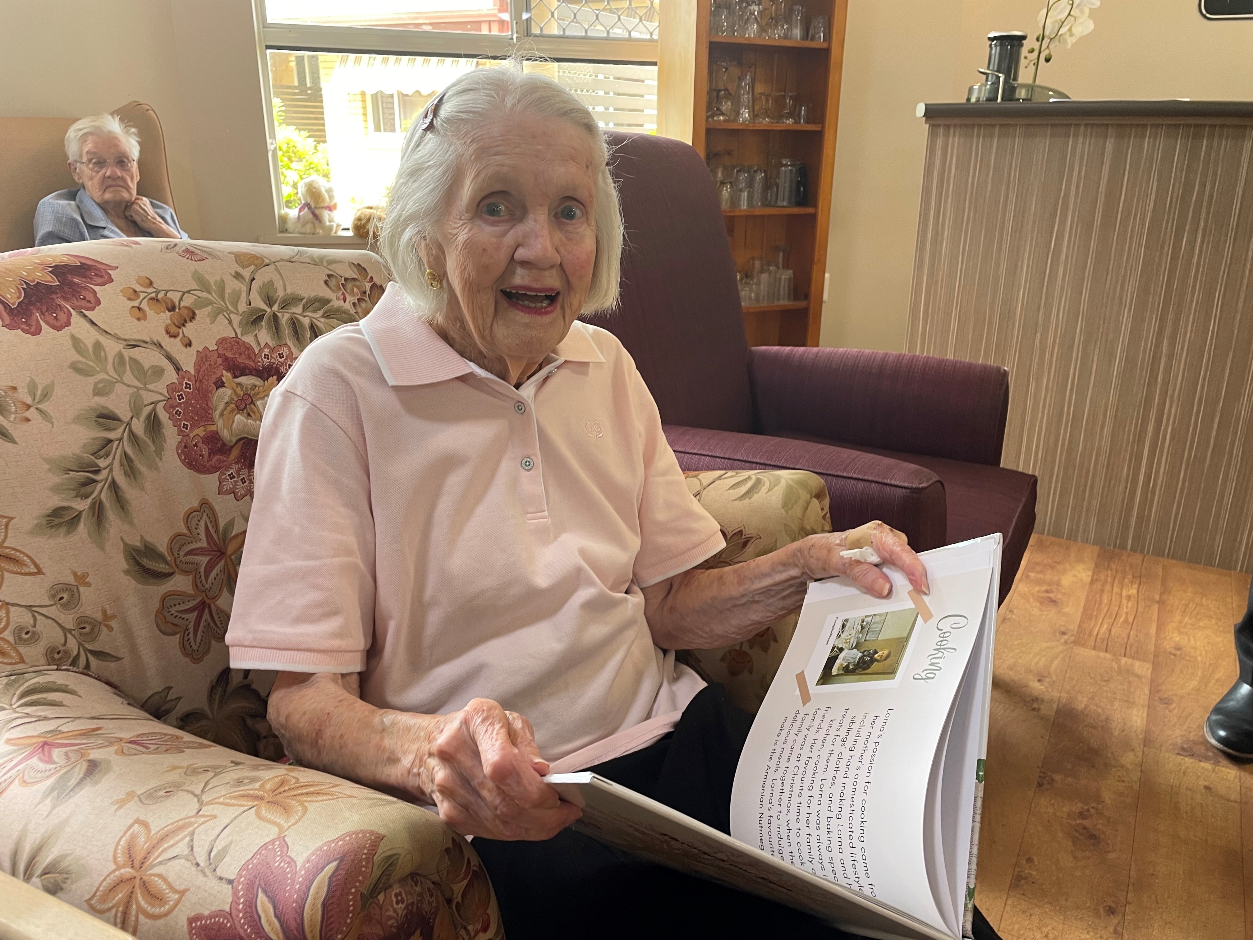An elderly woman sits on a lounge chair smiling as she looks through a book of memoirs.