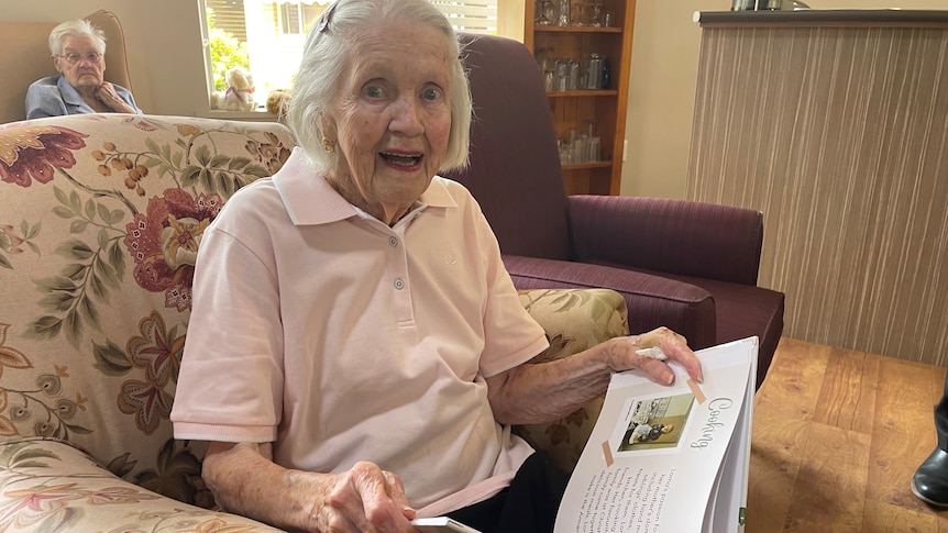An elderly woman sits on a lounge chair smiling as she looks through a book of memoirs.