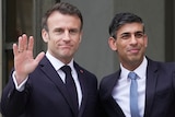 Macron, left and Sunak stand with an arm around one another. Macron lifts one hand in a wave to the press