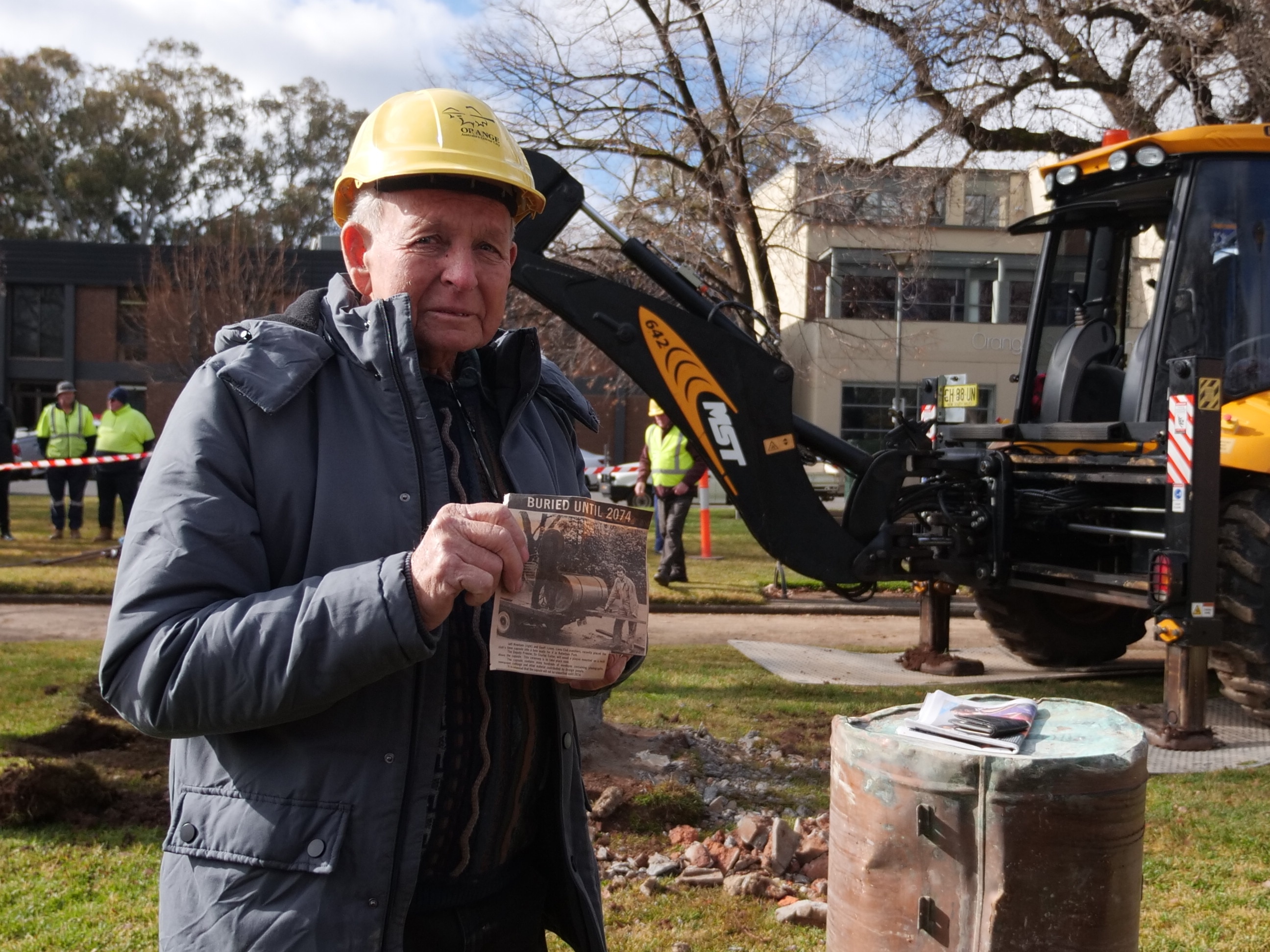 A man in a hard hat holding a newspaper clipping, standing in front of a digger and a metal drum. 