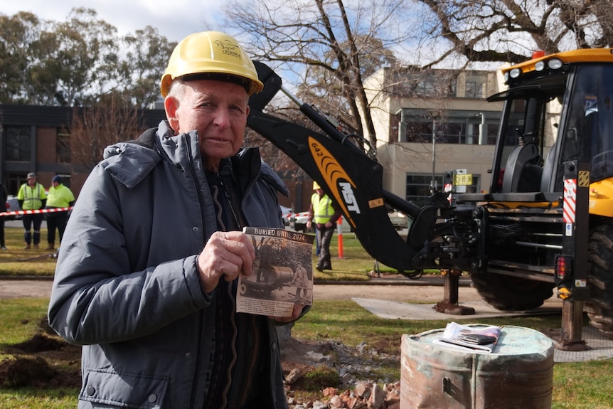 A man in a hard hat holding a newspaper clipping, standing in front of a digger and a metal drum. 