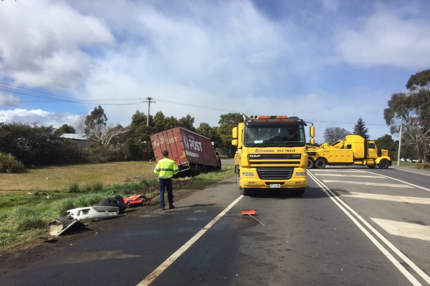 A yellow tow truck in the middle of the road, next to some debris from a crash. An Australia Post van is in the ditch