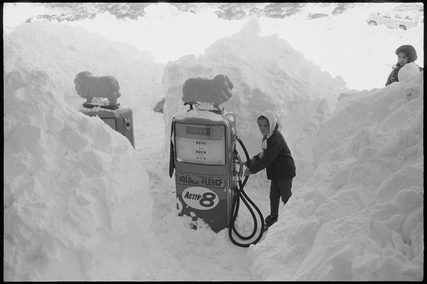 Black and white of old petrol bowser covered by snow and person trying to take the nozzle