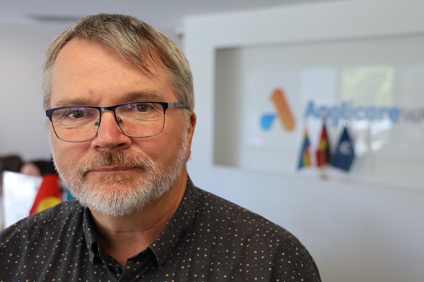 A serious man wears glasses, a dark shirt looks at the camera from the left side of the image, with an Anglicare sign behind.