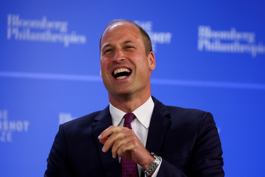 Prince William laughs. He is dressed in a suit. He's in front of a blue background and the words 'Bloomberg Philanthropies'.