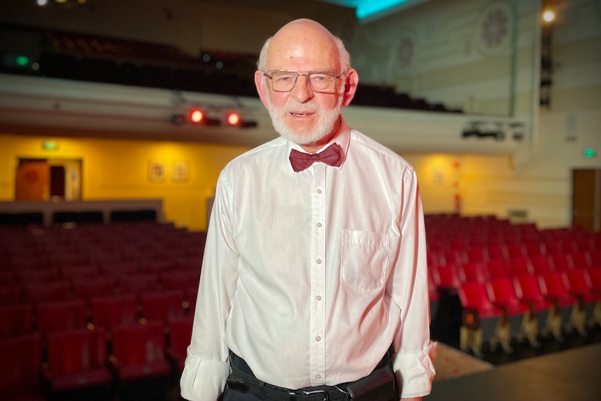 An older man in glasses, white shirt and red bow tie inside a theatre