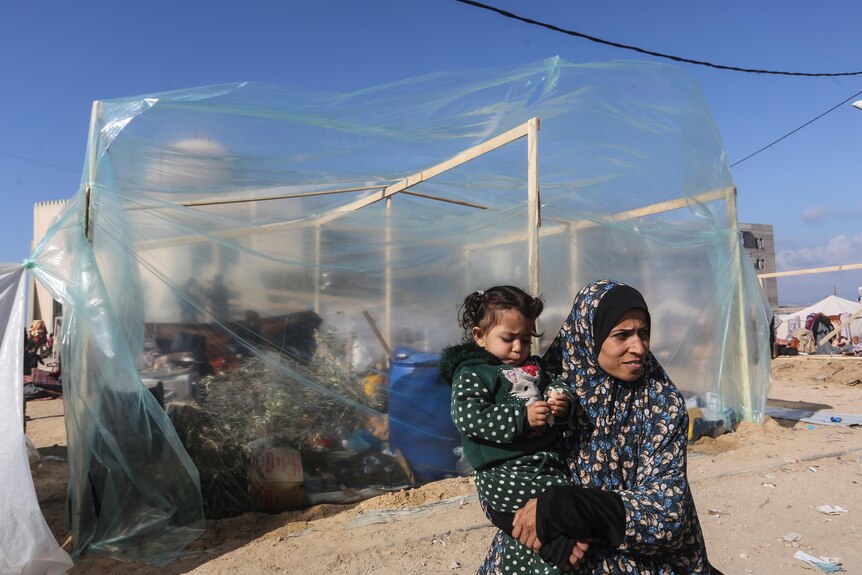 A woman in a hijab holding a young girl stands in front of a plastic, see-through tent
