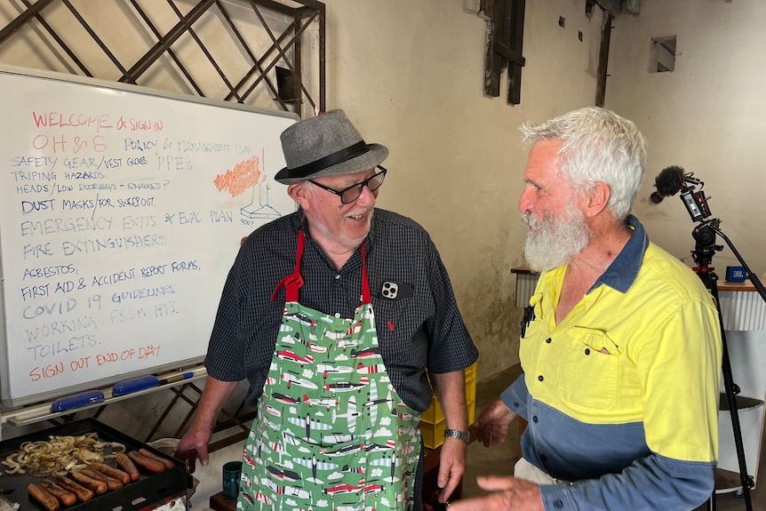 Two men chat in front of a whiteboard with a list of jobs while one cooks a barbecue.