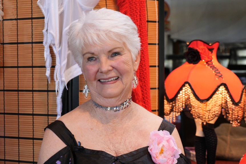 Woman in her 70s with short white hair, sparkly earrings and necklace, pink rose on her dress and lamp and feather boas behind.