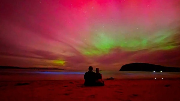Two people sit on beach with Aurora Australis in the sky