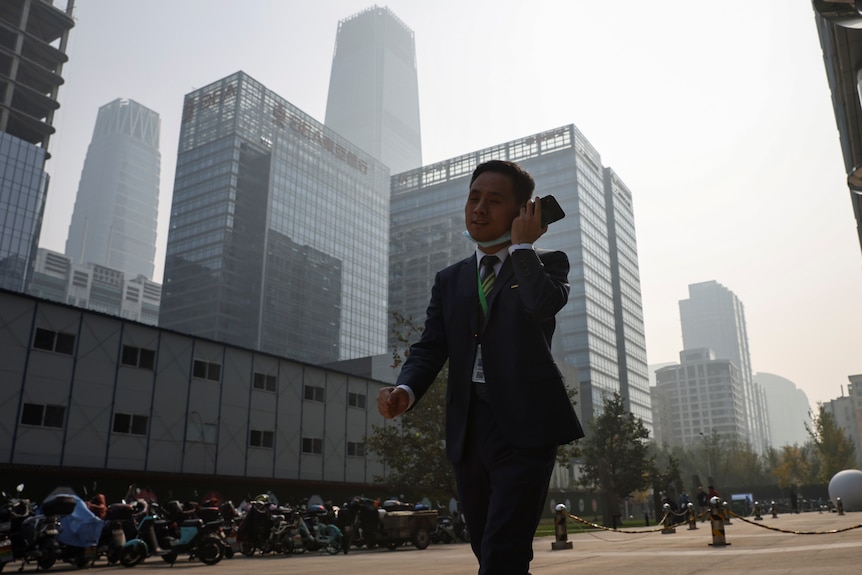 A man in a suit is pictured walking holding his phone up to his ear, he is surrounded by big grey buildings in Beijing CBD.