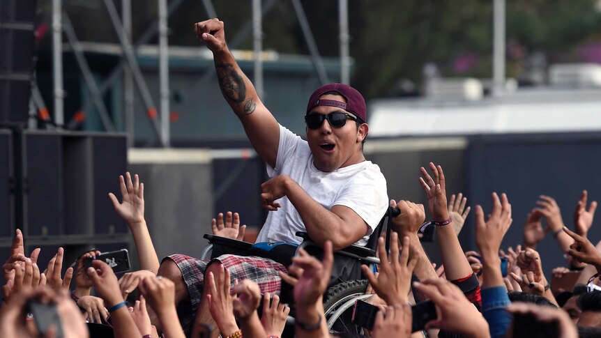 A young man in a wheelchair wearing a maroon hat pumps his fist along to music during a music festival