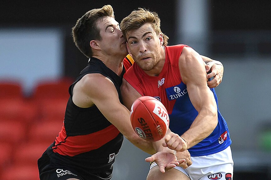 A Melbourne Demons AFL players handballs while being tackled around his upper body by an Essendon opponent on his right.