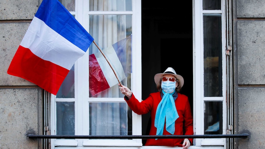 A woman in a face mask waves a French flag from her balcony