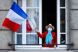 A woman in a face mask waves a French flag from her balcony