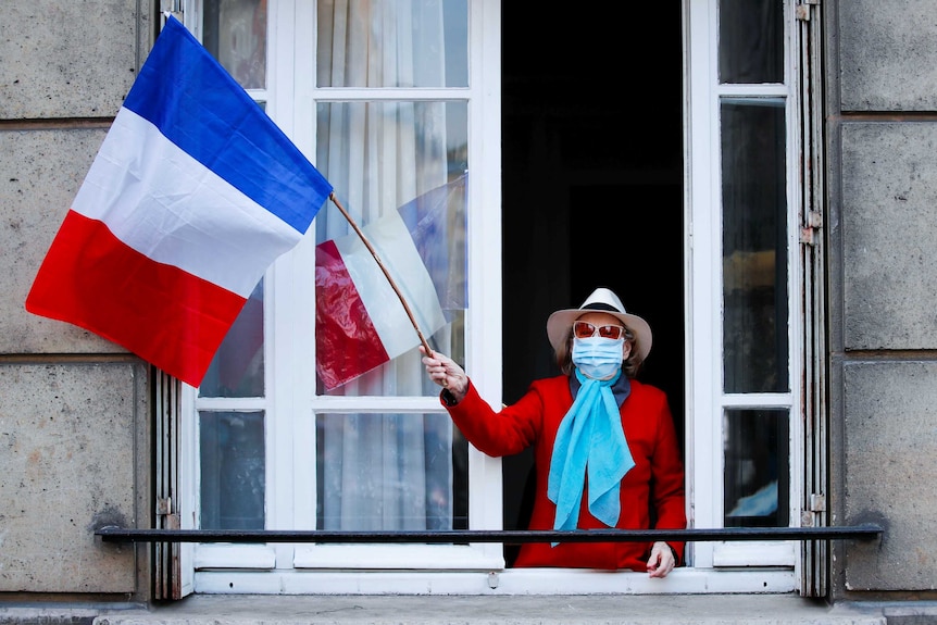 A woman in a face mask waves a French flag from her balcony