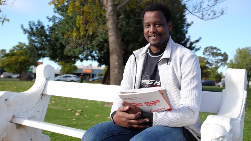 A man of African appearance sits on a white bench near a tree in a park. He is holding university textbooks.